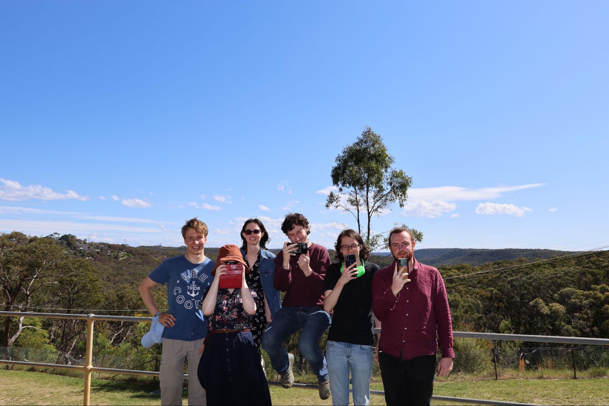 Group photo of six campgoers. From left to right: Sandro, Rena, Emma, Fergus, Jarod, Joel. All but Sandro and Emma are holding up their phone cameras. The landscape behind them is wooded and hilly.