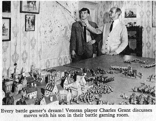 A black and white photograph of an older and younger man standing before a large table covered in models of soldiers in formation around a picturesque town. The caption reads, "Every battle gamer's dream! Veteran player Charles Grant discusses moves with his son in their battle gaming room."